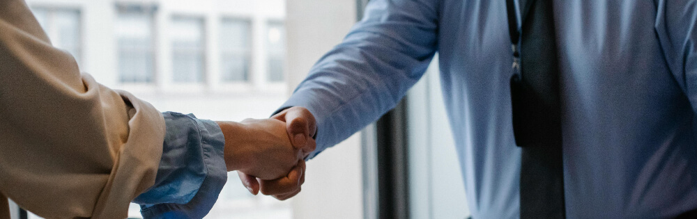 Zwei Personen im formellen Outfit schtteln sich die Hand, im Hintergrund ein Fenster mit verschwommener Aussicht.