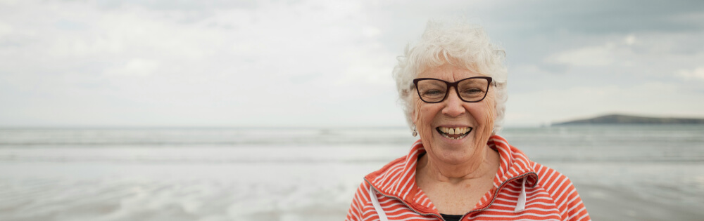 Eine ltere Frau mit weiem Haar und Brille lchelt frhlich am Strand. Sie trgt ein gestreiftes Oberteil und im Hintergrund ist das Meer mit einem wolkigen Himmel zu sehen.